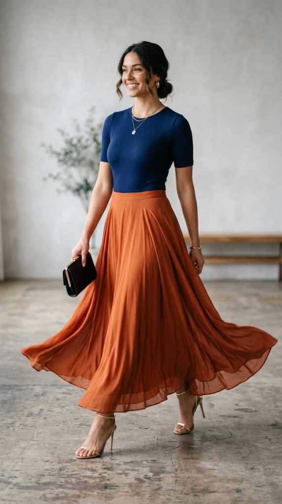 Woman walking indoors wearing a fitted navy blue short-sleeve bodysuit tucked into a flowing rust orange pleated maxi skirt, paired with gold strappy heels and a black clutch.